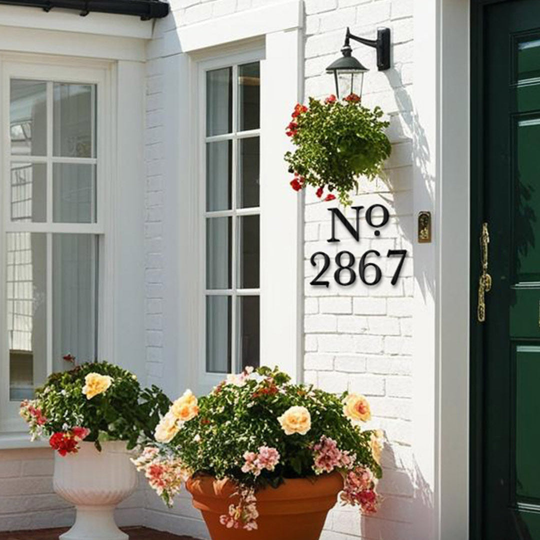House number with a "No" abbreviation in black metal '2867' on a white brick wall with potted flowers.