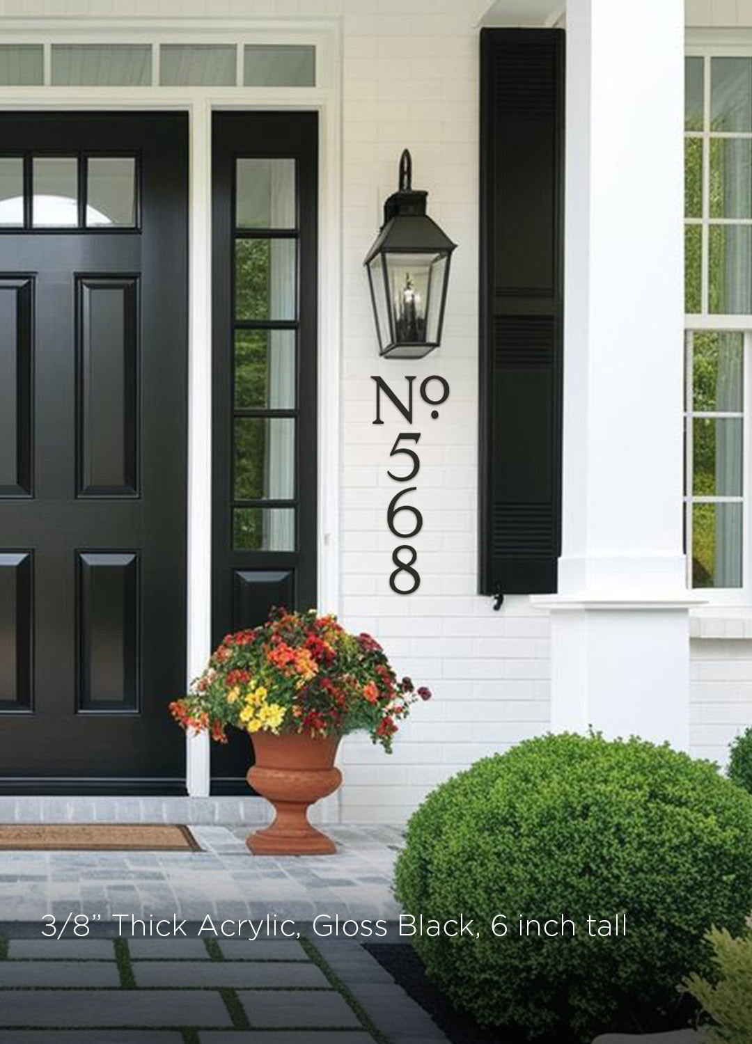 Black front door with decorative number plaque, flower pot, and lamp on a white house exterior.