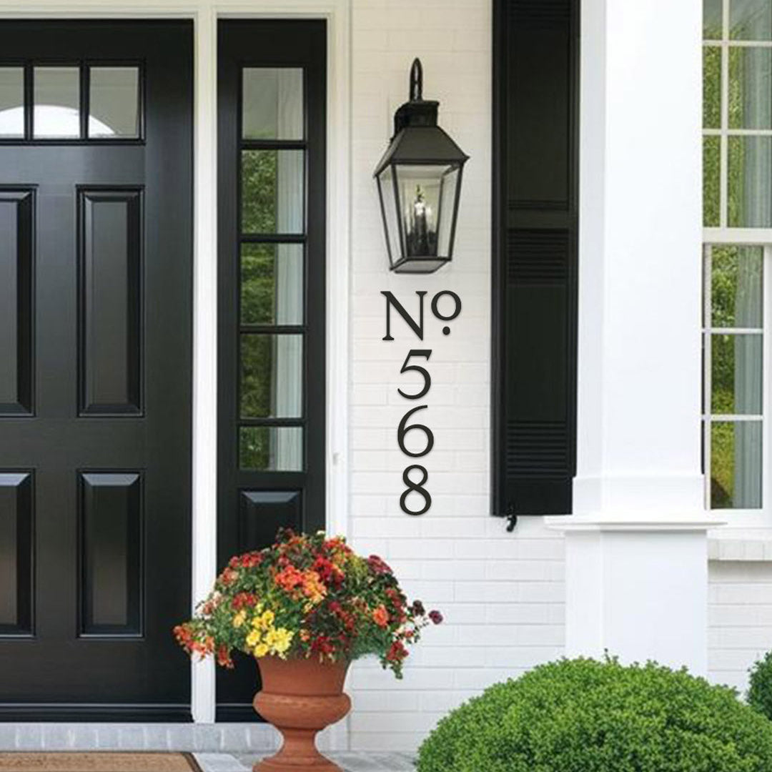 Black front door with decorative number plaque, flower pot, and lamp on a white house exterior.