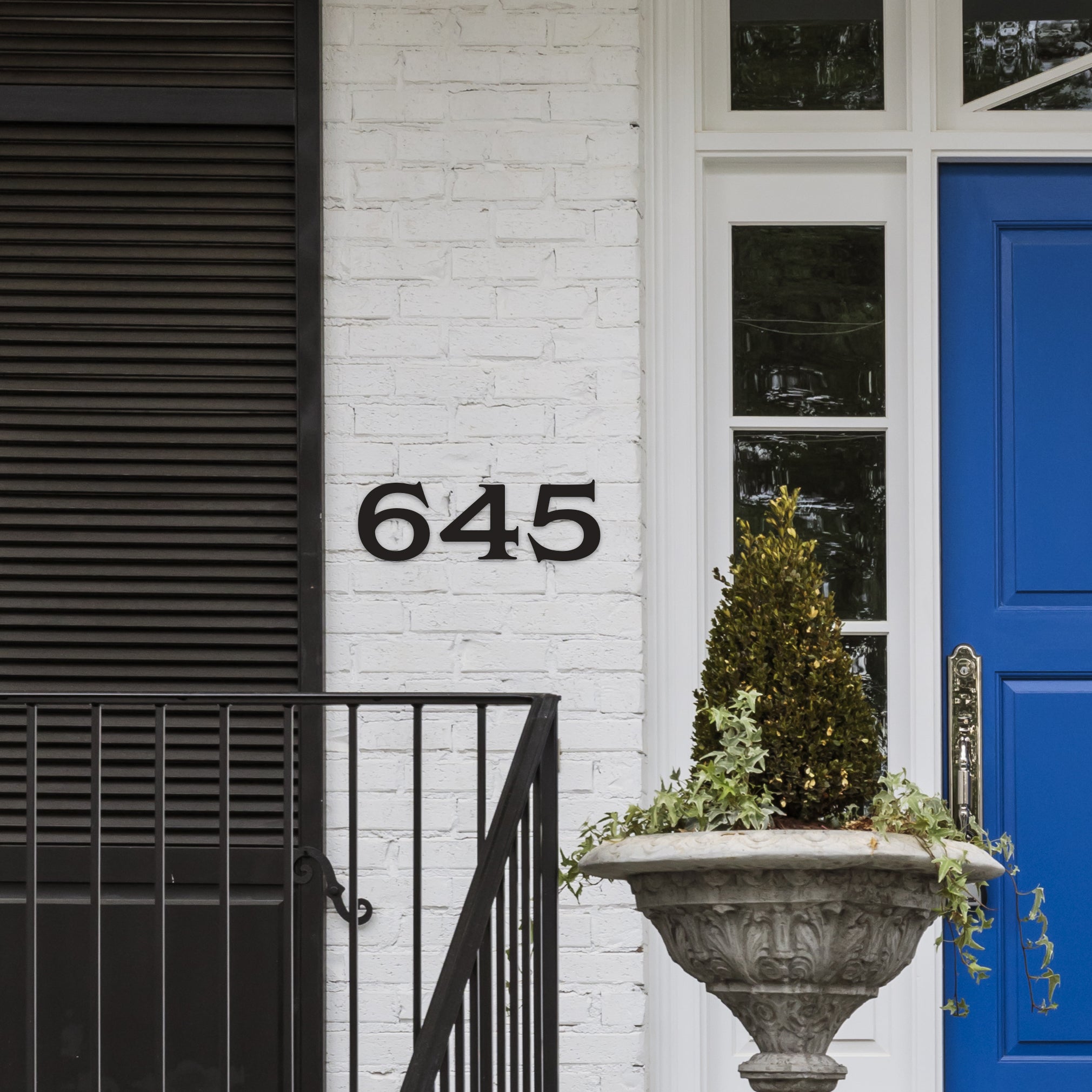 Black house numbers '645' mounted on a white brick wall above a blue door, flanked by a black shutter and a decorative plant pot.