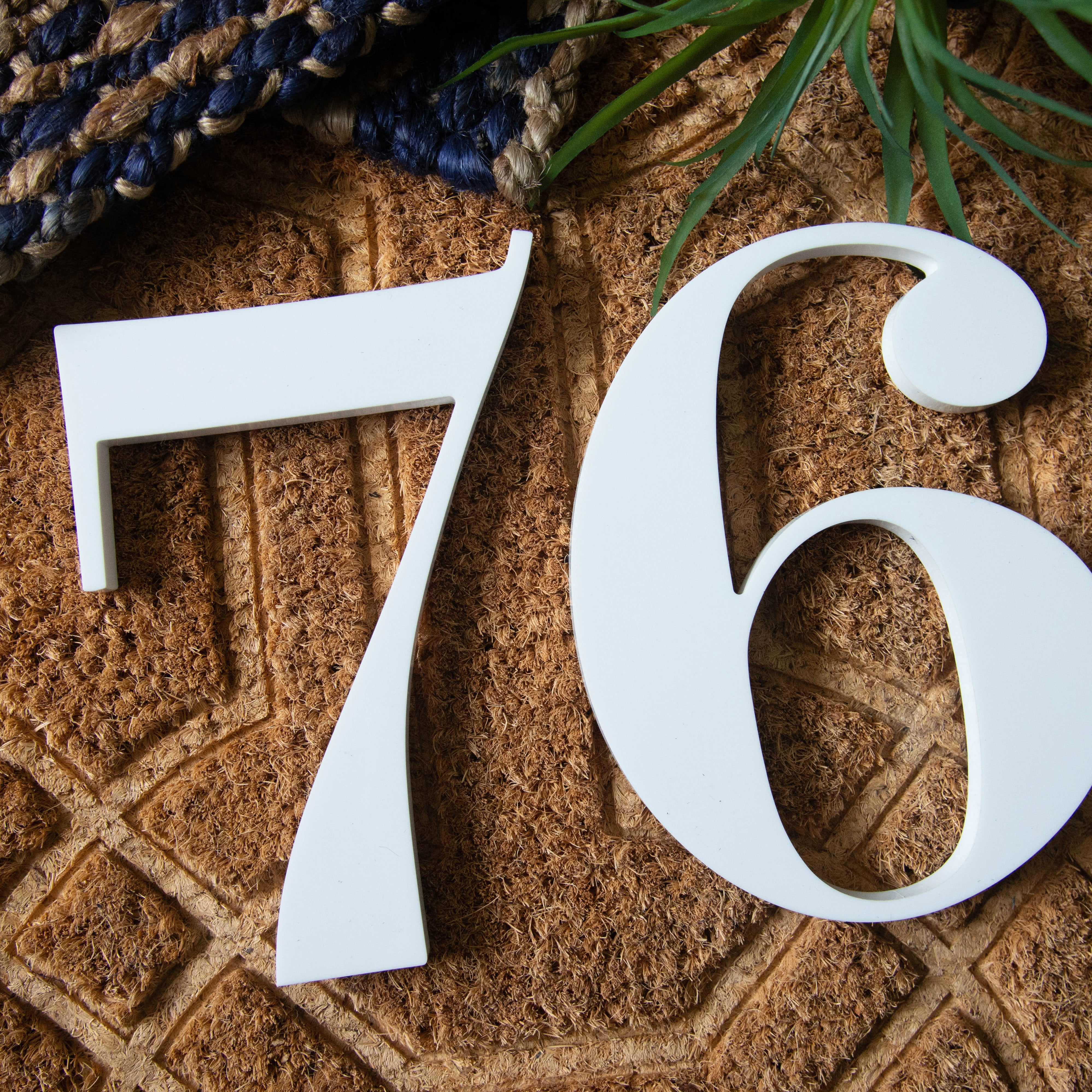 White house numbers '76' placed on a brown background with a partial view of a blue and white rug and some greenery.