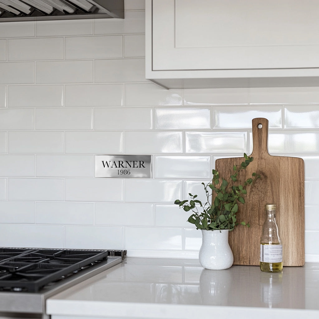 A polished Aluminum custom accent tile with the engraved text 'warner 1986' installed on a white tiled kitchen wall, next to a wooden cutting board and a small potted plant.