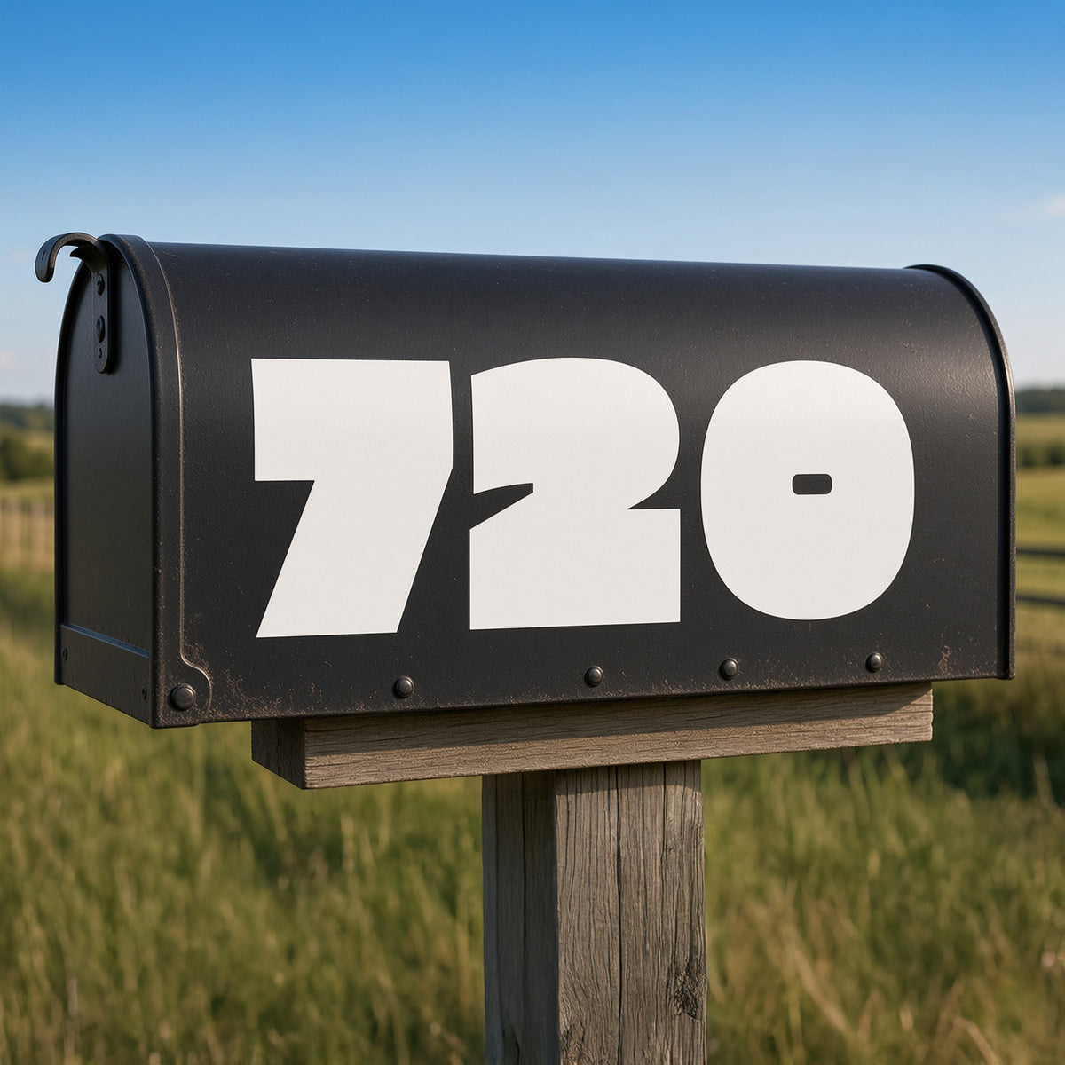 A black metal mailbox mounted on a wooden post in an outdoor grassy field under a blue sky, with large white numbers '720' printed on the front.