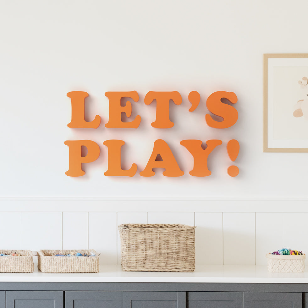The image shows a wall decor with the phrase 'LET'S PLAY!' written in bold, orange letters arranged on a wall shelf. The shelf is placed above a basket and below a framed picture.
