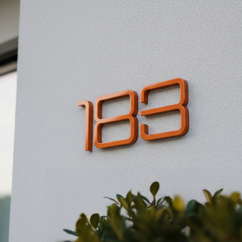 Orange house number '183' on a white wall with greenery at the bottom.