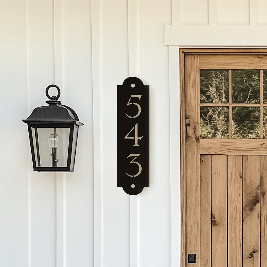 A black traditional style house number sign mounted on a white wall next to a wooden door, with a lantern-style light fixture to the left.