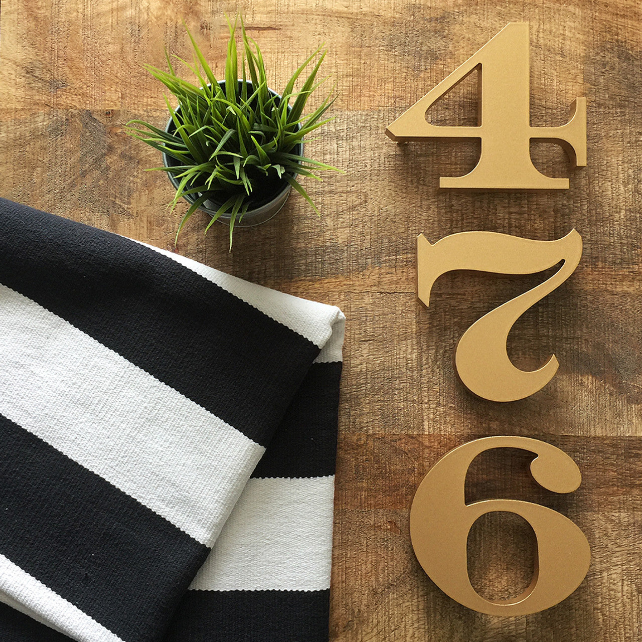 The image features two large, golden house numbers '4' and '7' placed on a wooden surface, with a small potted plant to the left and a striped fabric to the right, enhancing the visual appeal of the numbers.