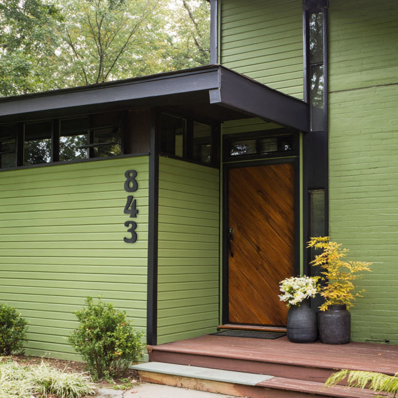 Green house with a wooden door and custom acrylic address number 843, surrounded by trees and plants.