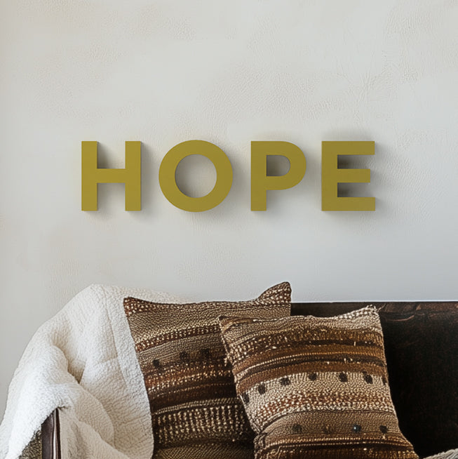 The image shows the word 'HOPE' written in capital letters on a plain wall, with each letter in a different color. The letter 'H' is in yellow, 'O' is in green, 'P' is in orange, and 'E' is in a shade of yellow-green, and the background is white with a shadow of the letters.