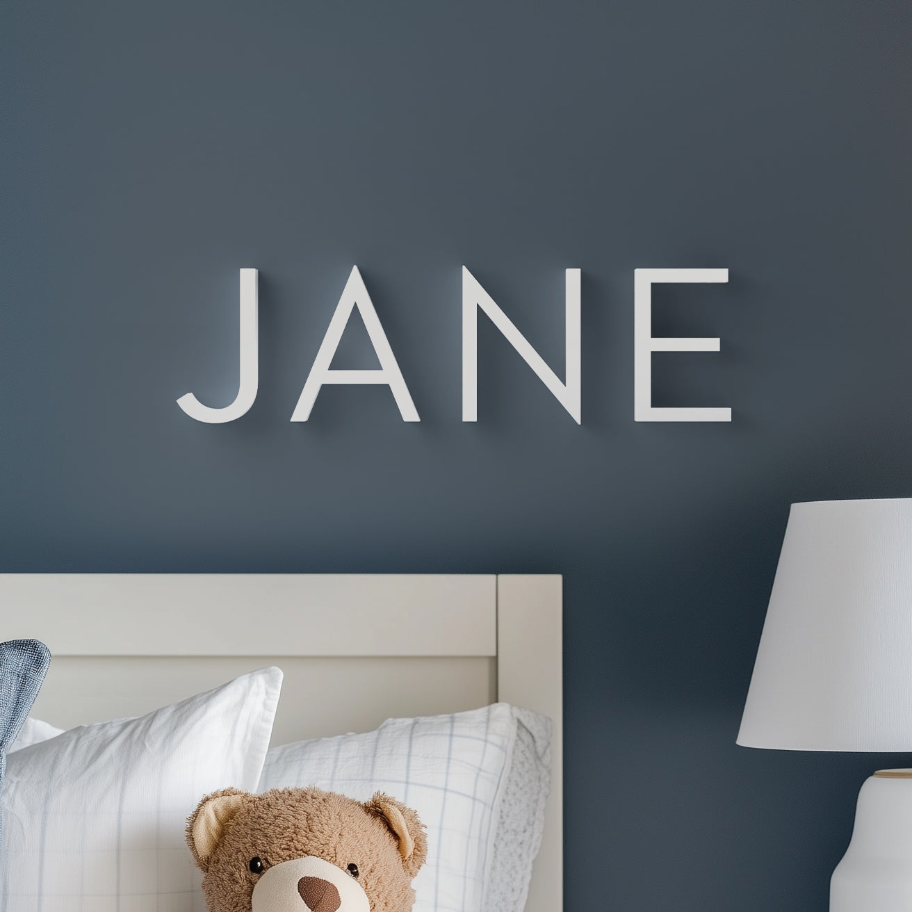 The image shows the wall decor product with the name 'JANE' in bold, white, upper-case letters on a dark blue wall above a bed with a teddy bear. The decor is used as a personalized touch to the room, suggesting a name for 'Jane'.