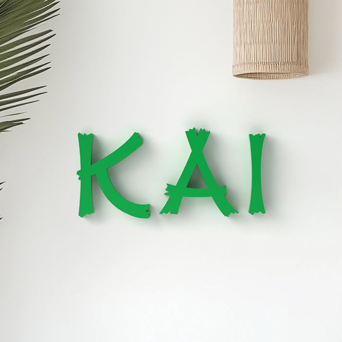 Custom personalized wall decor featuring the name 'KAI' in bold, jungle-inspired green lettering, displayed on a white wall with a palm leaf and decorative object to the side.
