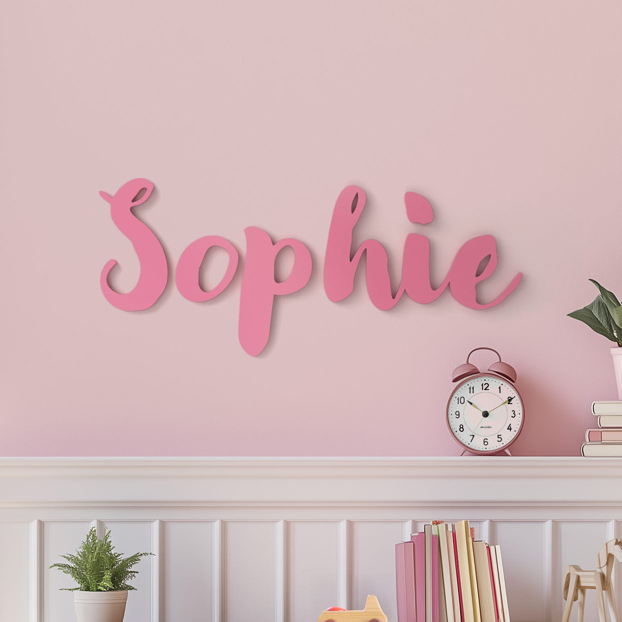 Customized wall decor spelling out the name Sophie in pink letters, mounted on a pink wall above a white shelf with books, a plant, and a clock.