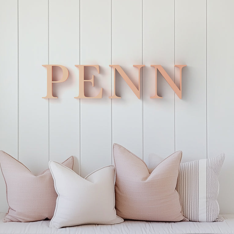 Acrylic letters spelling out the word 'PENN' mounted on a white wooden wall above a cushioned bench with neutral colored pillows.