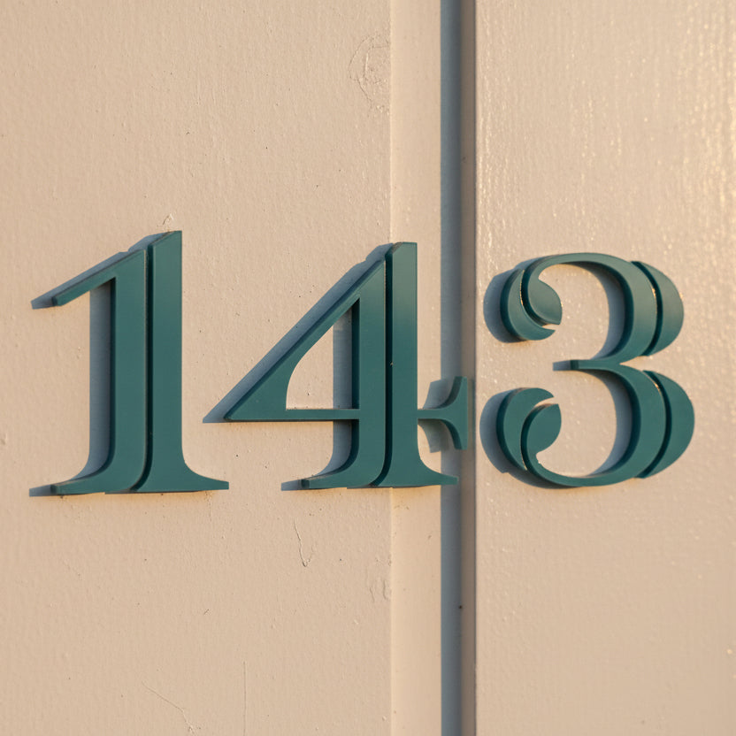 Traditional house numbers '143' in a dark teal color mounted on a beige stucco wall.