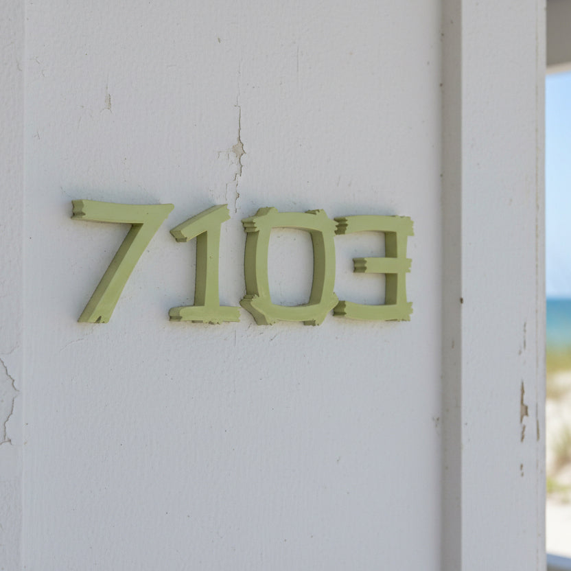 Green house numbers '7103' on a white wall with a beach view.