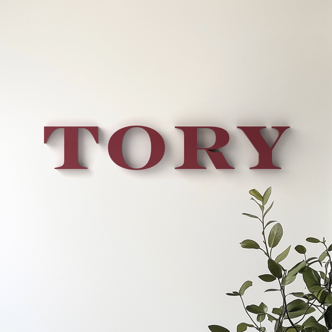 The image shows the word 'TORY' in bold, red, serif typography mounted on a white wall, with a green potted plant partially visible to the side.