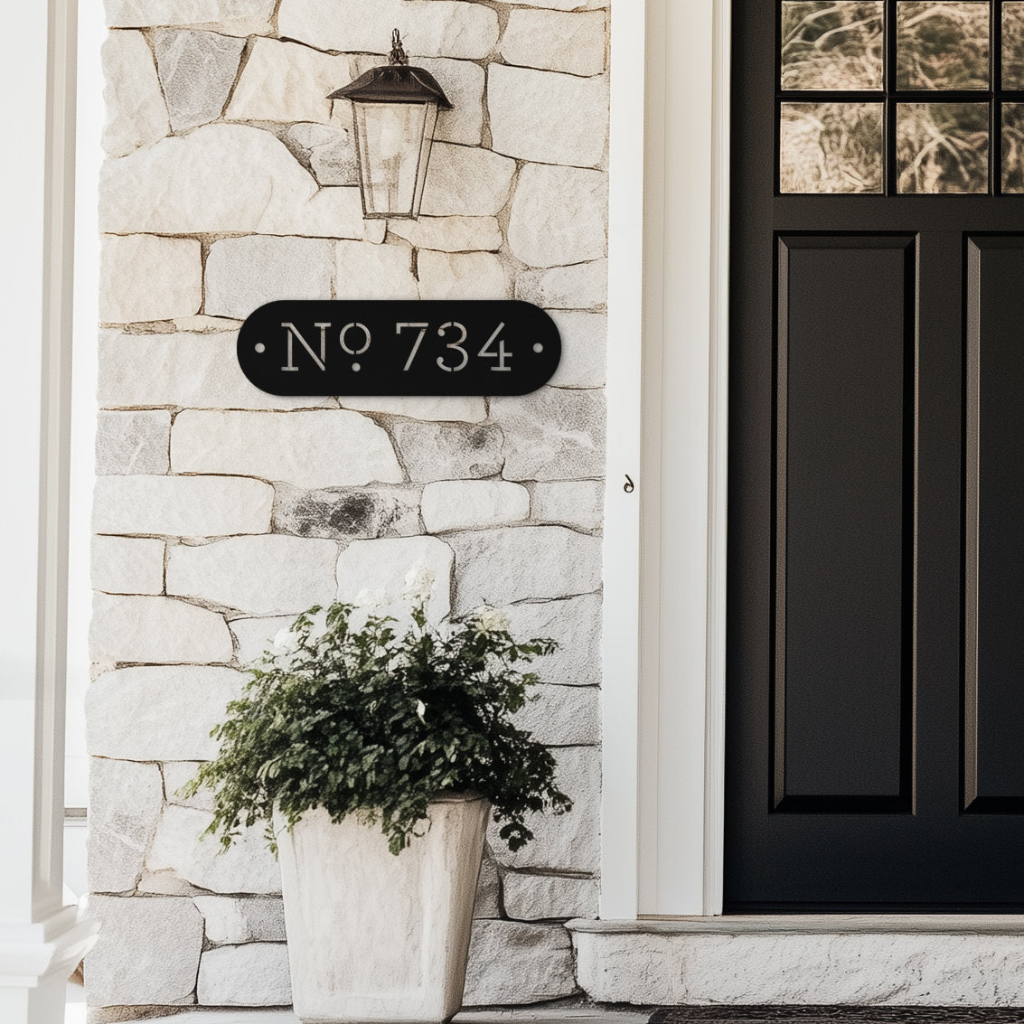 Modern farmhouse Black house number sign with the number 734 in white font, mounted on a white stone wall above a front door, next to a potted plant.