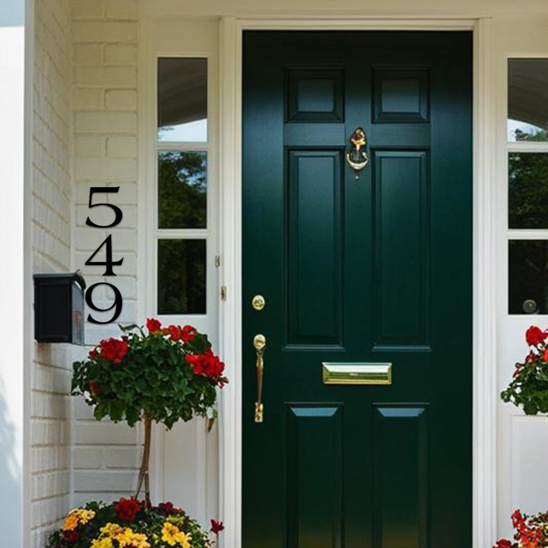 Green front door with gold handle and mail slot, flanked by potted plants on a white wall. Black metal 6 inch house numbers.