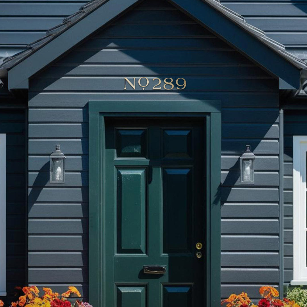 Dark blue house with a green door and decorative flowers in front with solid brass house numbers above door in a farmhouse style address numbers