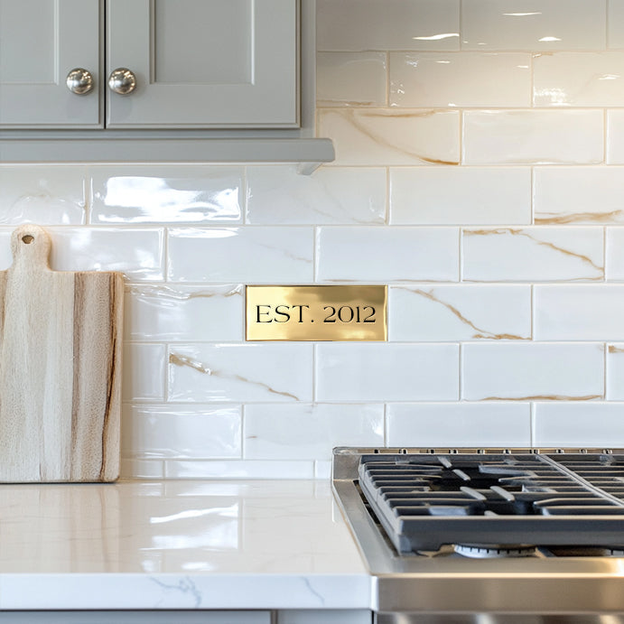 A custom accent tile with the inscription 'EST. 2012' installed on a kitchen wall behind the stove, surrounded by white marble tiles.
