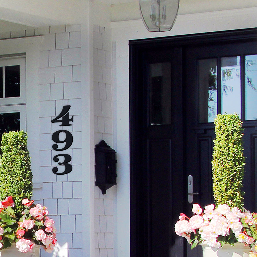 Black house numbers '493' mounted on a white wall above a black door, flanked by two potted plants with pink flowers.