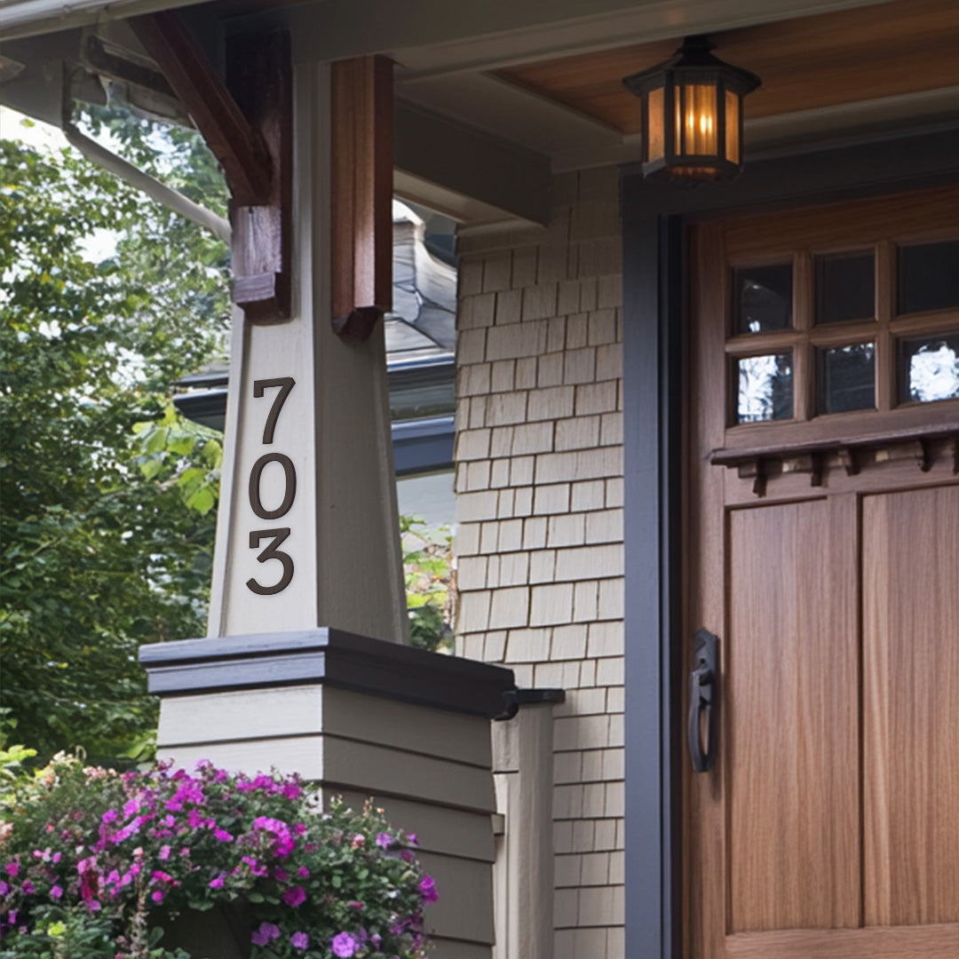 A close-up of a house number '703' mounted on a post beside a front door with a window, flanked by a floral arrangement and a lantern.