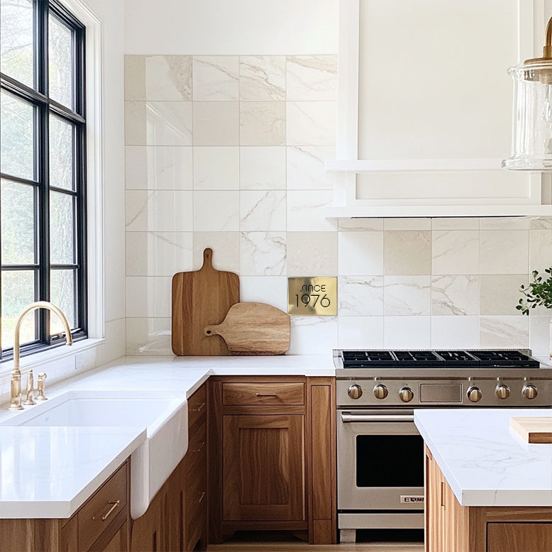 A kitchen setting featuring custom brass accent tiles with a marble backsplash, wooden cabinets, and a stainless steel range. Tile reads "since 1976"