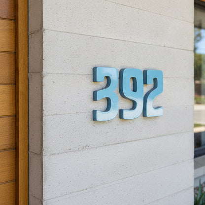 3D house numbers '392' in a light blue metallic finish mounted on a beige textured stucco wall next to a wooden door frame.