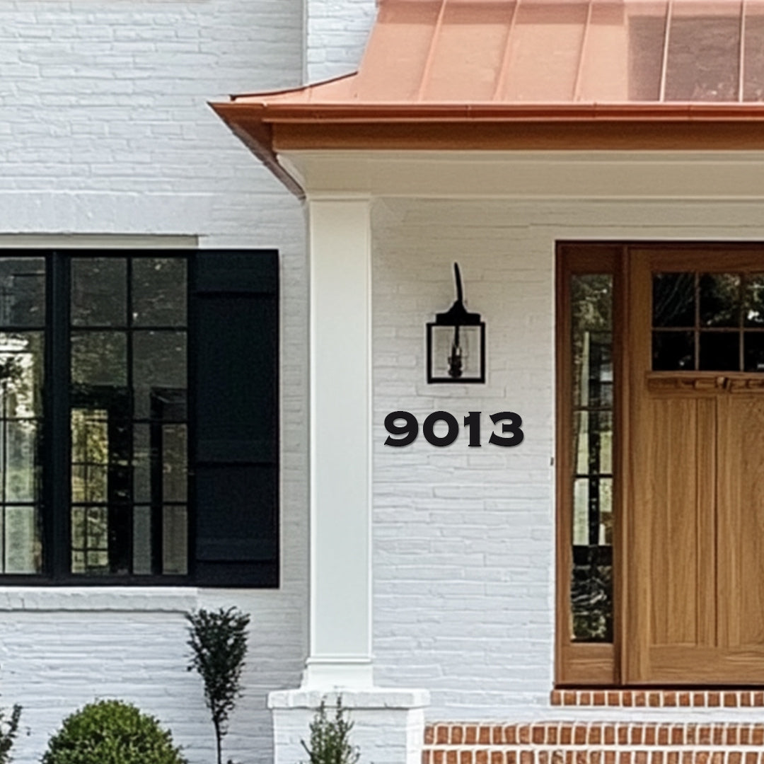White house with black window frames and a wooden front door, featuring black metal house numbers '9013' mounted on the wall. Copper roof
