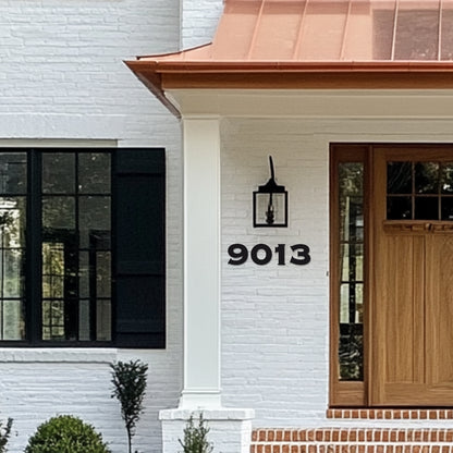 White house with black window frames and a wooden front door, featuring black metal house numbers '9013' mounted on the wall. Copper roof