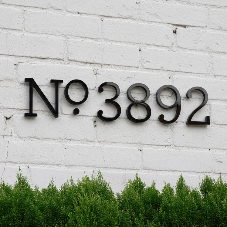 Black numbers '3892' on a white brick wall with greenery at the bottom.