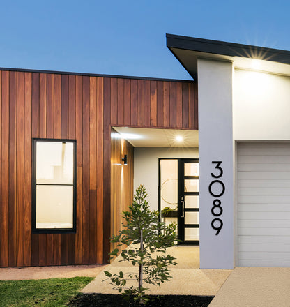 Modern house numbers '3089' mounted on a white column with a wooden wall in the background, under a blue sky.