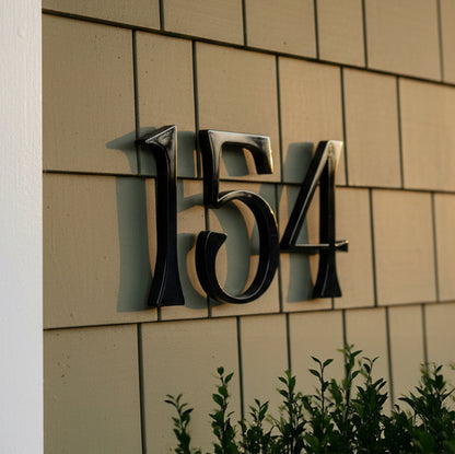 Black house numbers '154' on a beige tiled wall with greenery at the bottom.