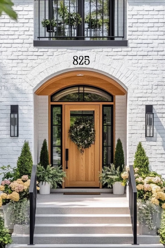 Wooden front door with wreath, white brick wall, and decorative plants. House numbers on brick arch in a decorative style
