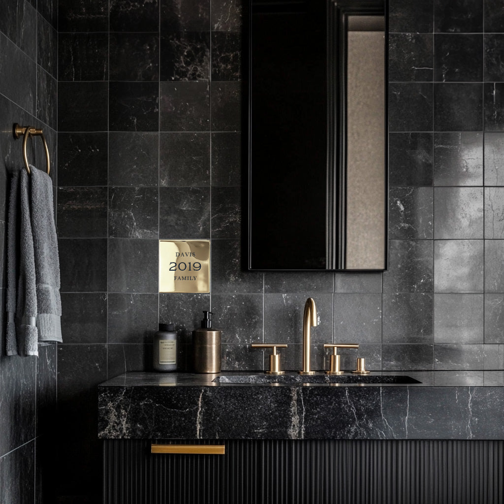 A black tiled bathroom wall featuring a custom brass accent plaque grouted into the tile with the text 'Davis 2019 family' displayed in a traditional style font.