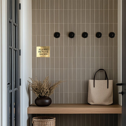 A custom brass accent tile with the text 'Please remove shoes' mounted on a wall above a wooden bench, next to a basket and a potted plant in a tiled mudroom entryway