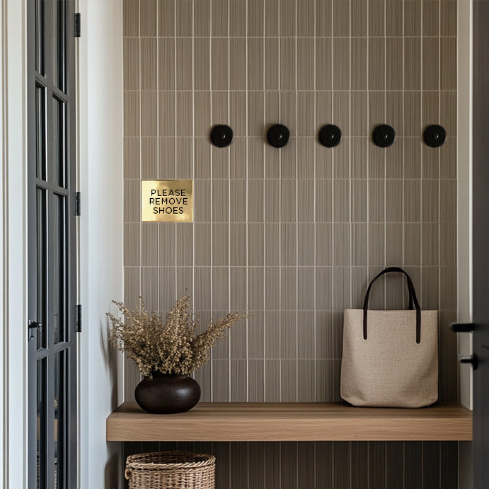 A custom brass accent tile with the text 'Please remove shoes' mounted on a wall above a wooden bench, next to a basket and a potted plant in a tiled mudroom entryway