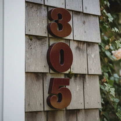 Rust-colored house numbers '305' on a wooden wall with greenery in the background