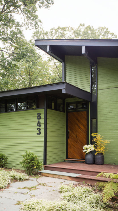 Green house with a wooden door and custom acrylic address number 843, surrounded by trees and plants.