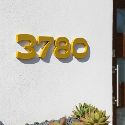 Gold house numbers '3780' on a white wall with a plant in the foreground.