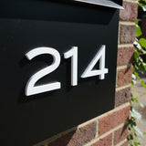 White dimensional numbers 214 mounted on black metal mailbox against red brick wall. Shown in: White