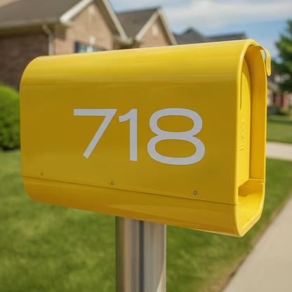 A bright yellow mailbox mounted on a metal post in a suburban front yard, with the white number 718 printed on the front and a blurred house, grass, and sidewalk in the background.