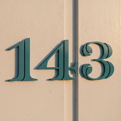 Traditional house numbers '143' in a dark teal color mounted on a beige stucco wall.