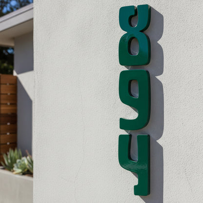 Dark green 3D metal house numbers reading '894' mounted vertically on a textured white stucco wall.