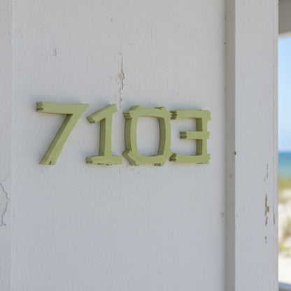 Green house numbers '7103' on a white wall with a beach view.