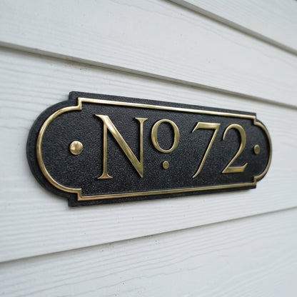 Decorative house number plaque displaying No. 72 in raised brass letters on black textured background mounted on white siding. Shown in: Brass · Black background