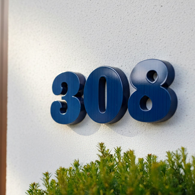 Blue house number '308' on a white wall with greenery at the bottom.